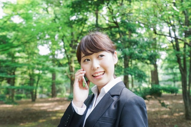 恵まれた自然の中でスーツを着た女性が笑顔で電話をかけるシーン(イメージ図)(画像はphotoACより)